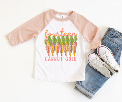 a white raglan with peach sleeves and a graphic with fourteen different colored carrots and a the words fourteen carrot gold the word fourteen is above the carrots in a pink cursive with a yellow shadow and the words carrot gold are underneath the carrots