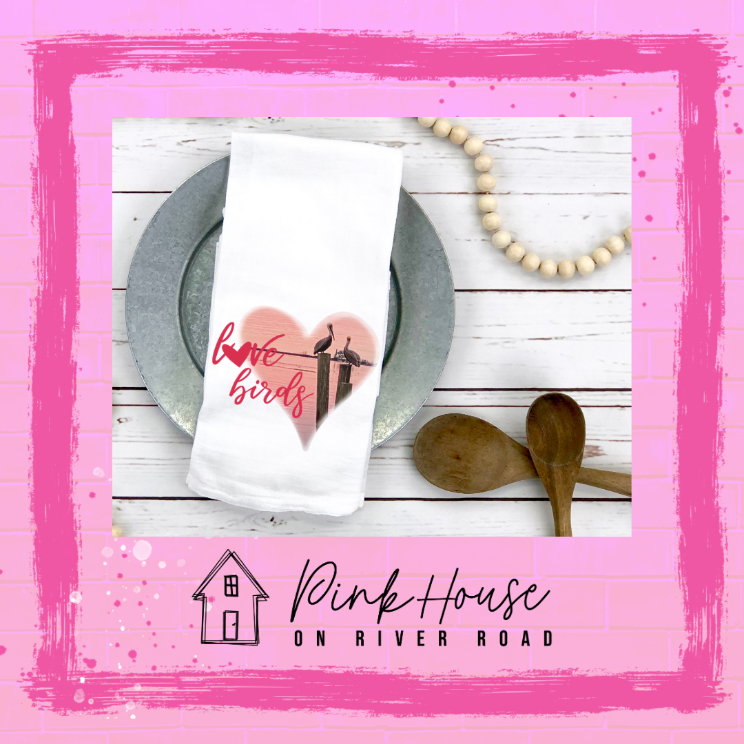 A white Dish Towel with graphic. Graphic is a pink heart with a photo of some pelicans standing on post in the water. To the left of the heart are the words love birds in a pink cursive font and the O in love has been replaced with a heart.