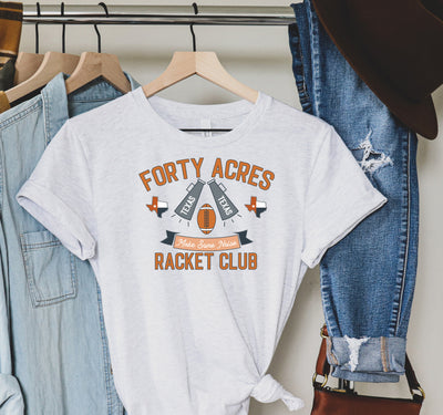 Heather White Tshirt on hanger.  Tshirt has art that says ""Forty Acres Racket Club" the art also has a banner that says make some noise with a football, two megaphones that say Texas and two Texas Graphics.