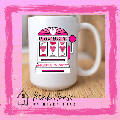 A white coffee mug with a graphic. Graphic is of a white slot machine with pink accents at the top of the slot machine is an arch with different shades of pink and a heart cut out of the middle. Underneath the are are the words Love machine in hot pink with white dots to look like lights. the reels of the slot machines show three geometric hearts. The slot pull has a pink base with a white handle and pink knob on it. Under the reels are the words Jackpot Winner in pink. 