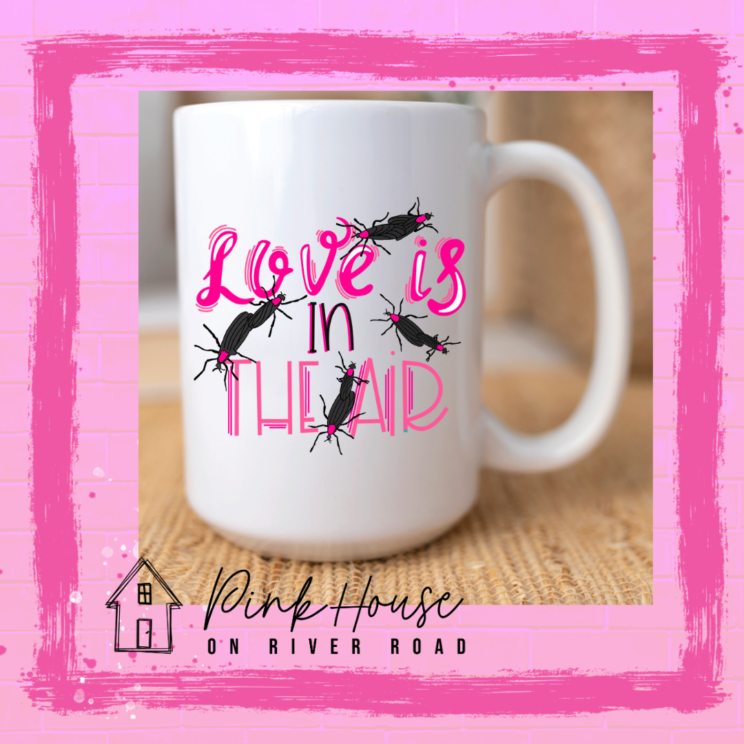 White Mug with Graphic. Graphic says " Love Is in the Air". "Love Is" is a hot pink cursive font with white highlights and hot pink accent lines underneath is the word "In" Is it in a black font with a hot pink highlight. on the bottom is "The Air" in Light pink with light pink accent lines. There are also black flying bugs with hot pink spots behind their heads