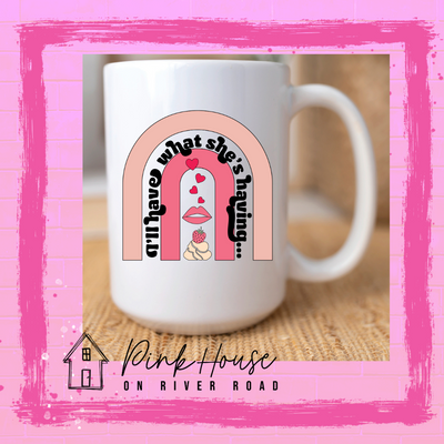 A white coffee mug with a strawberry dessert and a set of lips with hearts, there is a pink arch going over the art. There is a retro font arched over the pink art that says "I'll have what she's having..." in black with another arch above that in light pink.