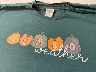 A long sleeve stonewash blue tee with a fall graphic. There are five different colored pumpkins each with a different letter that spells out the word gumbo and underneath the word weather in a white cursive font