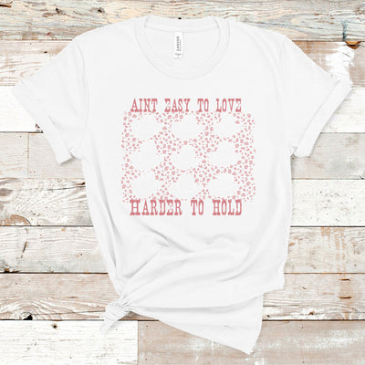white tee with graphic. Graphic is pink weathered letters at the top that read " Aint Easy To Love" underneath the text there are 9 different styles of cowboy hats outlined in pink with a pink leopard print in between the cowboy hats, text below the hats reads " Harder To Hold"