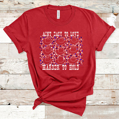 Red tee with graphic. Graphic is white weathered letters at the top that read " Aint Easy To Love" underneath the text there are 9 different styles of cowboy hats outlined in white with a white and blue leopard print in between the cowboy hats, text below the hats reads " Harder To Hold"