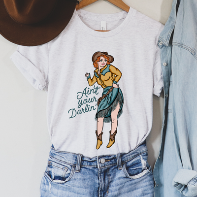 Ash Tee. Art is a red haired pin up cowgirl with a blue fringe skirt and a yellow pearl snap with blue accent with brown boots, hat, and belt. Rope font that reads Ain't your Darlin'