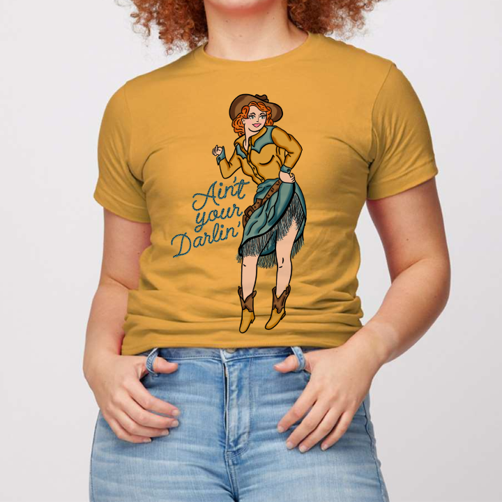 Mustard Yellow Tee. Art is a red haired pin up cowgirl with a blue fringe skirt and a yellow pearl snap with blue accent with brown boots, hat, and belt. Rope font that reads Ain't your Darlin'