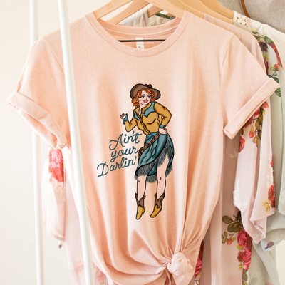 Peach Tee.  Art is a red haired pin up cowgirl with a blue fringe skirt and a yellow pearl snap with blue accent with brown boots, hat, and belt. Rope font that reads Ain't your Darlin'
