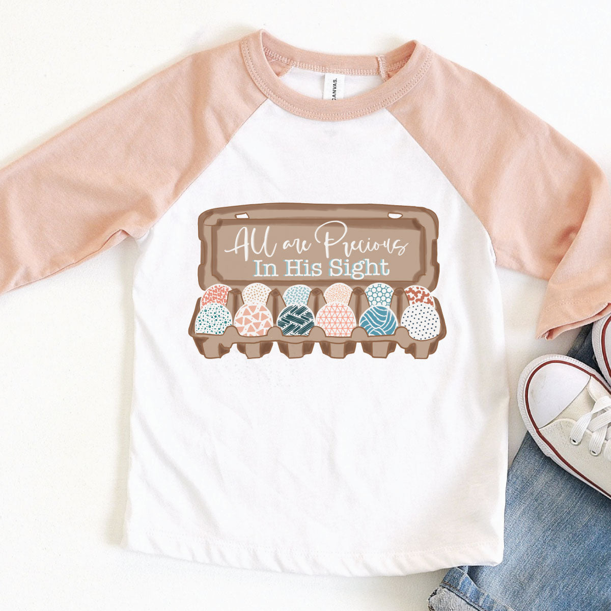 White with Peach Sleeves Raglan Shirt. Graphic is a carton of Easter eggs all with a different patterned designed and on the lid of the open carton read All are precious in his sight.