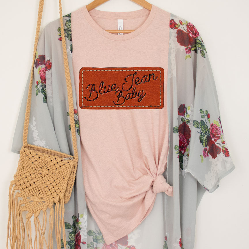 Peach tee has a graphic that looks like a leather patch sewn onto the shirt and the words Blue Jean Baby in rope font on the patch. Pictured under a floral kimono 