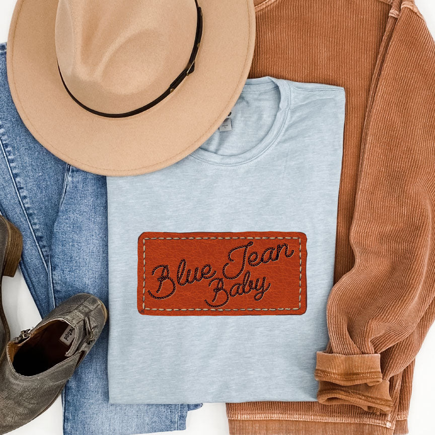 Stonewash Denim Tee has a graphic that looks like a leather patch sewn onto the shirt and the words Blue Jean Baby in rope font on the patch. Pictured with Jeans a hat and tan sweatshirt