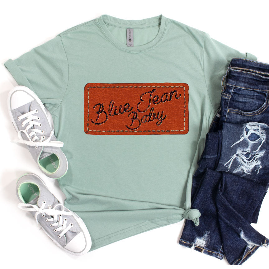 Stonewash green tee with a graphic that looks like a leather patch sewn onto the shirt and the words Blue Jean Baby in rope font on the patch. Pictured with a paid of grey converse and a pair of distressed jeans.