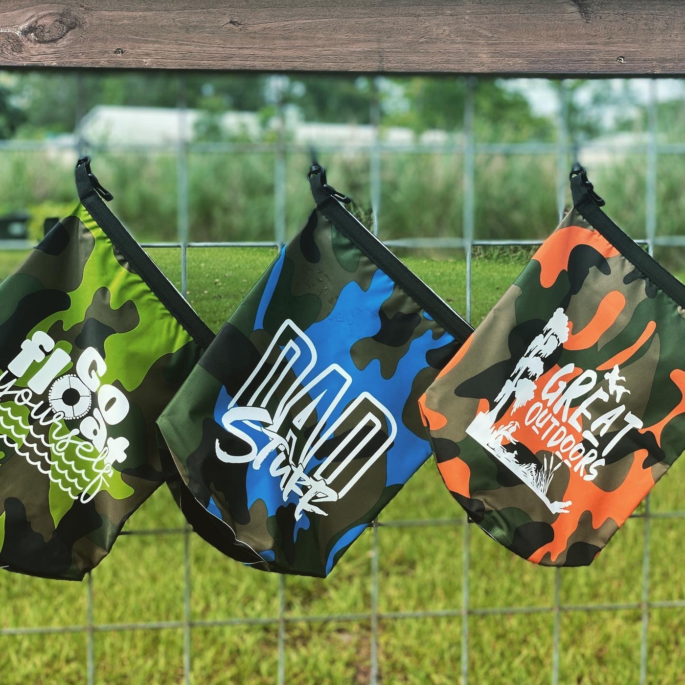 Three camo dry bags hanging on a fence. From left to right there is a green camo bag that says " Go Float Yourself" The O in float is a floatie and there are waves, In the middle is a blue camo dry bag that says dad stuff, and on the far right is an orange camo bag that says great outdoors and has ducks trees and reeds