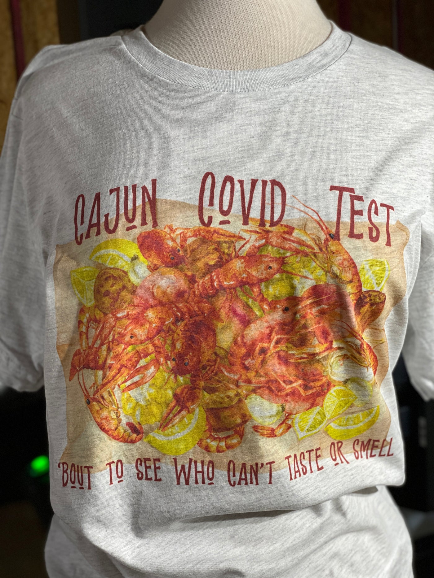 Ash Tee with Graphic. Graphic is a piece of craft paper loaded with crawfish, sausage, potatoes, corn, onions, garlic and lemons. Text above graphic says "Cajun Covid Test" and the text below says "Bout To see who can't taste or smell" All text in red
