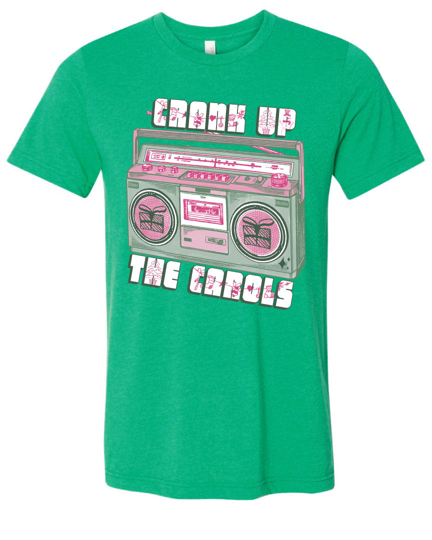 Green tee with graphic. Graphic has an old school boom box in the middle, the boom box is green with pink highlights and the speakers have the outline of presents on them. There is text above the boombox that is white with a green shadow and pink Christmas designs that says "Crank up" and below the boom box that says "The Carols"