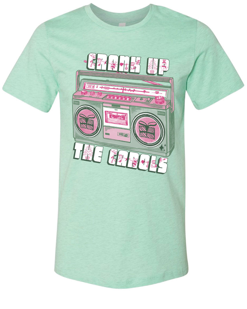 Light Green tee with graphic. Graphic has an old school boom box in the middle, the boom box is green with pink highlights and the speakers have the outline of presents on them. There is text above the boombox that is white with a green shadow and pink Christmas designs that says "Crank up" and below the boom box that says "The Carols"