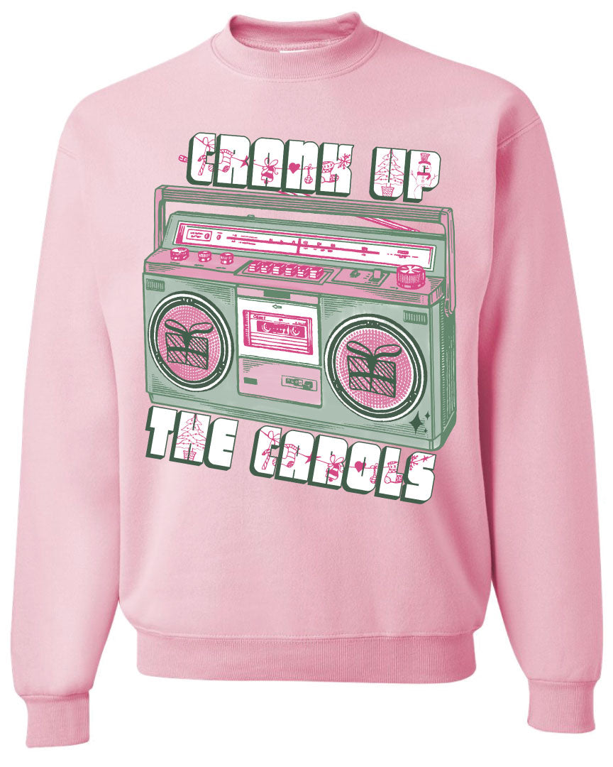 light pink sweatshirt with graphic. Graphic has an old school boom box in the middle, the boom box is green with pink highlights and the speakers have the outline of presents on them. There is text above the boombox that is white with a green shadow and pink Christmas designs that says "Crank up" and below the boom box that says "The Carols"