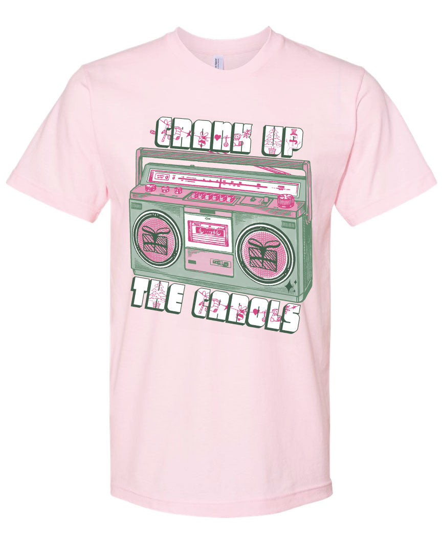 Pink tee with graphic. Graphic has an old school boom box in the middle, the boom box is green with pink highlights and the speakers have the outline of presents on them. There is text above the boombox that is white with a green shadow and pink Christmas designs that says "Crank up" and below the boom box that says "The Carols"