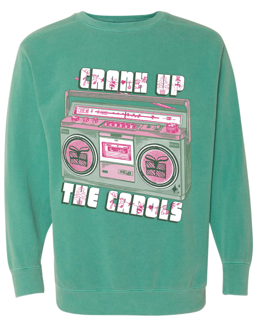 Green sweatshirt with graphic. Graphic has an old school boom box in the middle, the boom box is green with pink highlights and the speakers have the outline of presents on them. There is text above the boombox that is white with a green shadow and pink Christmas designs that says "Crank up" and below the boom box that says "The Carols"