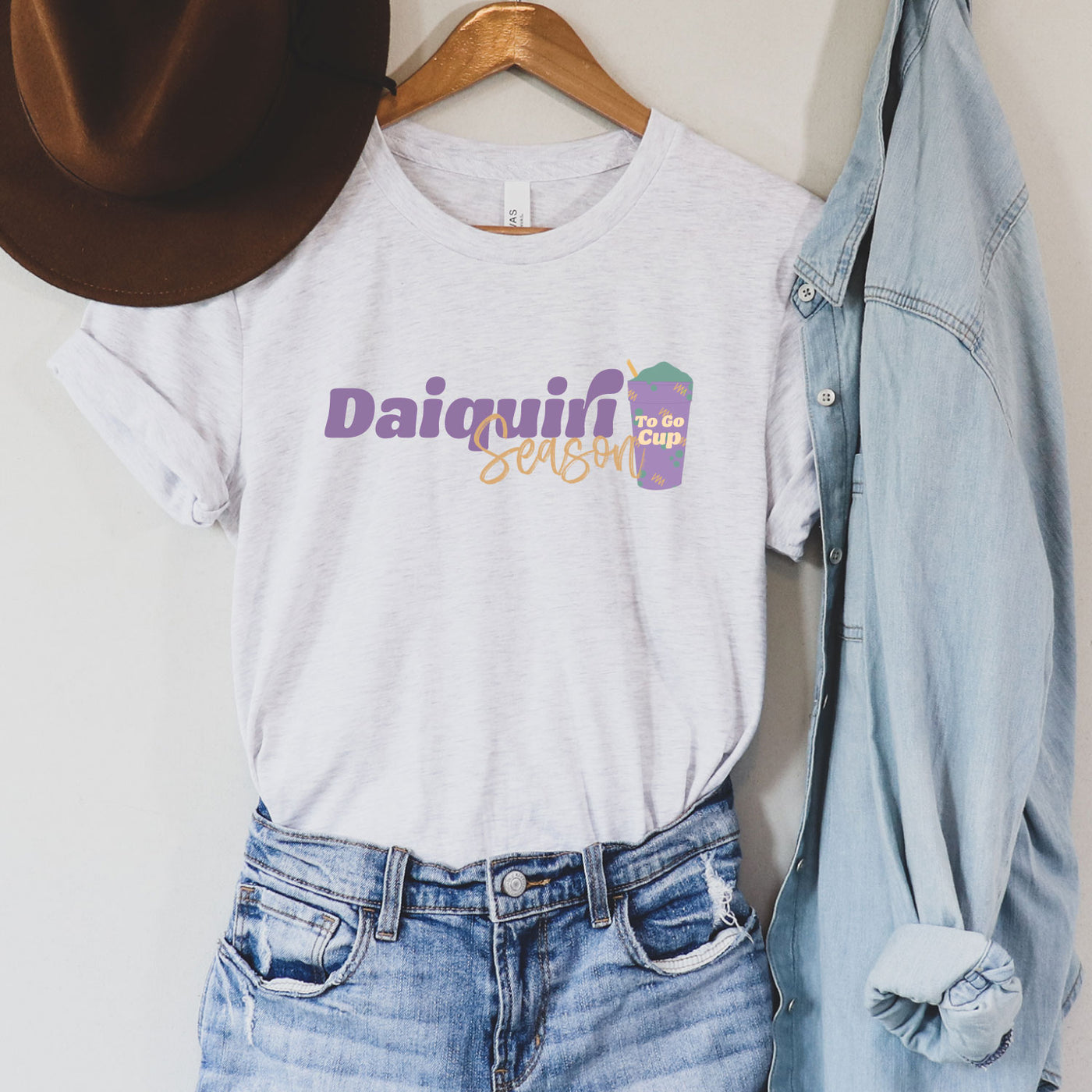 Ash gray tee with graphic. Graphic says Daiquiri Season, Daquiri is in purple letters and season is underneath it in a smaller yellow cursive font. To the right of the text there is a purple cup with green liquid and a yellow straw, the cup has yellow and green designs and the words To Go Cup in yellow.