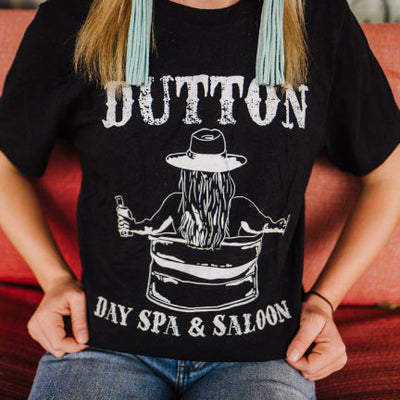 Blonde woman in blue jeans a black shirt with a white graphic on it. The graphic Has the word Dutton in old western letters at the top and Day Spa & Saloon at the bottom, in between the words you can see a womans back with long hair under her cowboy hat sitting in a water trough holding a bottle of whiskey.