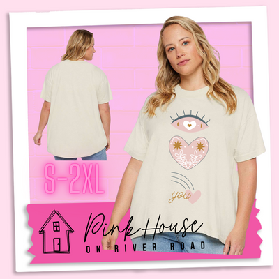 A blonde woman in a cream oversized HiLo tee with the graphic of an eye with a heart for the pupil and underneath that a heart with a floral and star design and a shooting heart with the word you. The graphic represents eye heart you