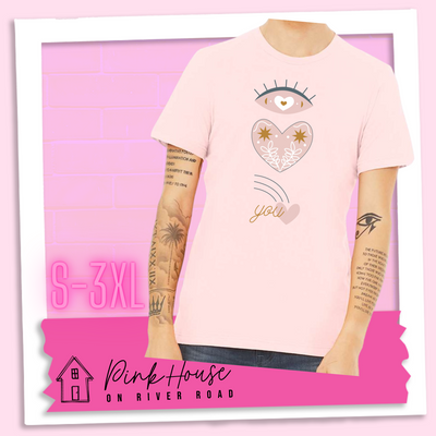 Soft Pink tee with a graphic of an eye with a heart for the pupil and underneath that a heart with a floral and star design and a shooting heart with the word you. The graphic represents eye heart you