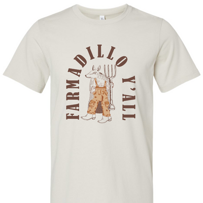 Dust tee with a graphic of an armadillo wearing tan overalls and a pair of boots holding a pitch fork. the words "Farmadillo Y'all" arching around the graphic.