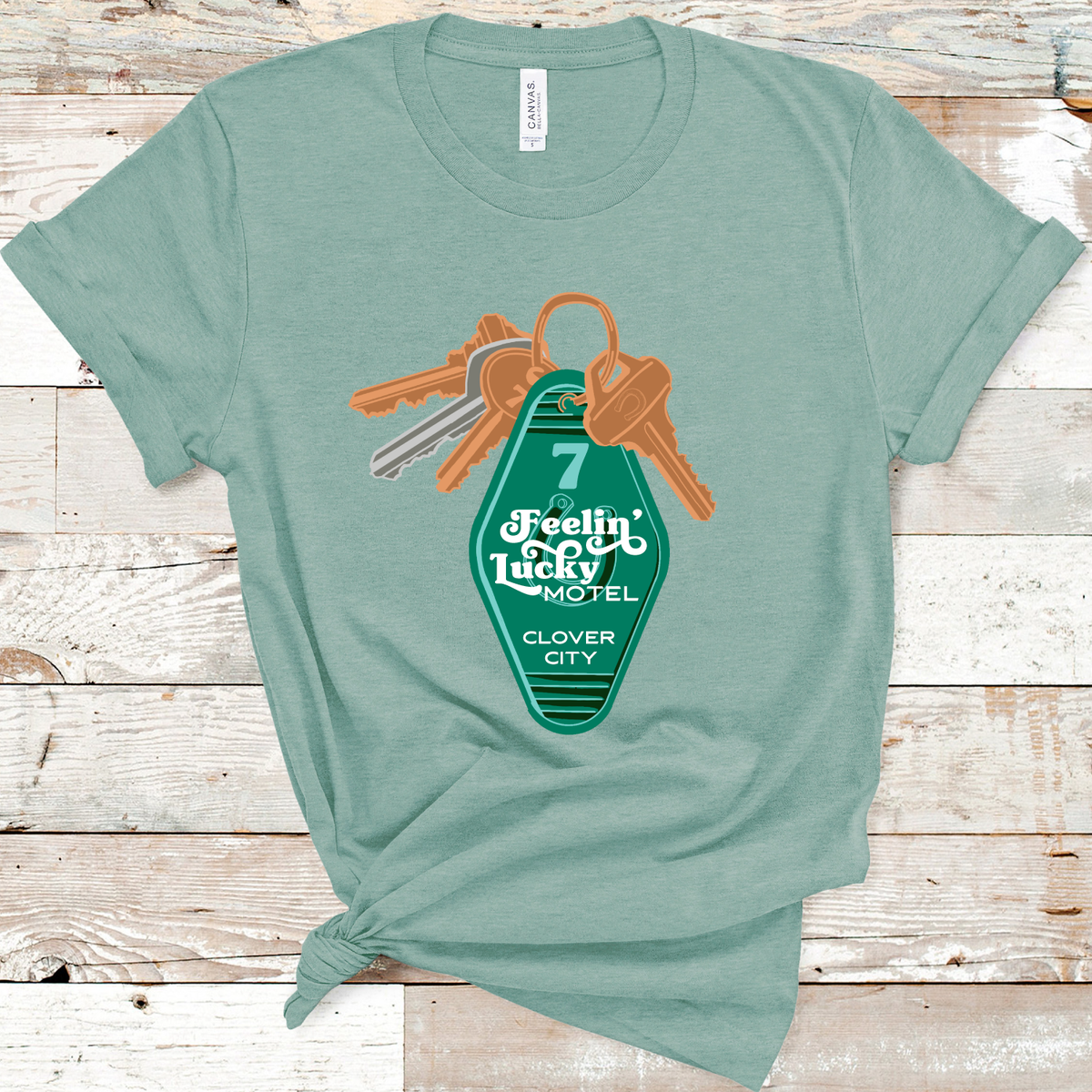Dusty Blue Tee. A set of keys with a vintage motel key chain. Key chain is green with a number 7 and a horse shoe. There is a retro white font over the horse shoe that says Feelin' Lucky Motel with Clover City underneath. 