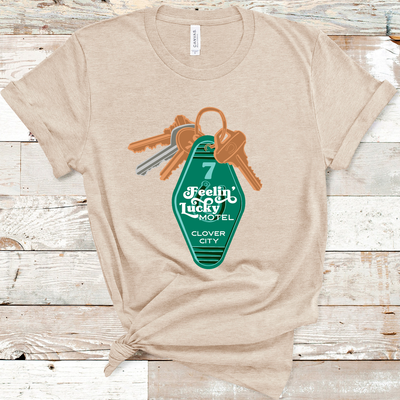 Heather Tan Tee. A set of keys with a vintage motel key chain. Key chain is green with a number 7 and a horse shoe. There is a retro white font over the horse shoe that says Feelin' Lucky Motel with Clover City underneath.