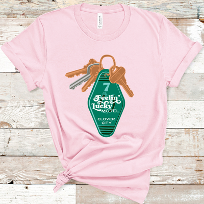 Light Pink Tee. A set of keys with a vintage motel key chain. Key chain is green with a number 7 and a horse shoe. There is a retro white font over the horse shoe that says Feelin' Lucky Motel with Clover City underneath.