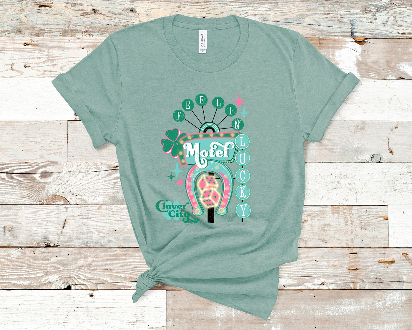 Dusty Blue Tee. Graphic of a vintage Neon Sign The word feeling with each letter in green circles at the top with the word lucky down the side with each letter in a turquoise circle. There is a sign in the center with the word motel in it, underneath there is a horse shoe with dice inside at he bottom of the sign. There is also a four leaf clover with the word clover city over it in green.