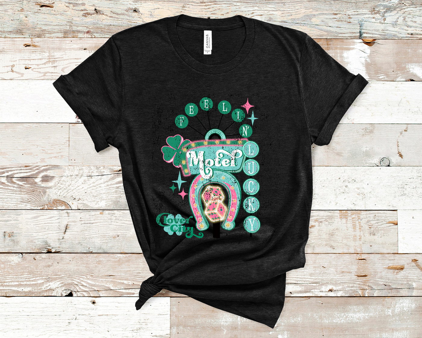 Heather Black Tee. Graphic of a vintage Neon Sign The word feeling with each letter in green circles at the top with the word lucky down the side with each letter in a turquoise circle. There is a sign in the center with the word motel in it, underneath there is a horse shoe with dice inside at he bottom of the sign. There is also a four leaf clover with the word clover city over it in green.