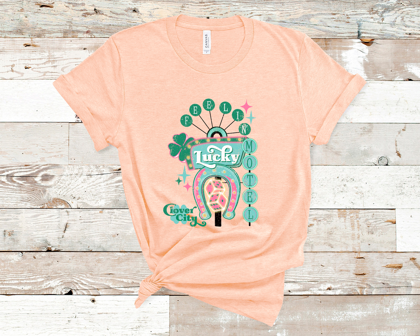 Heather Peach Tee. Graphic of a vintage Neon Sign The word feeling with each letter in green circles at the top with the word lucky down the side with each letter in a turquoise circle. There is a sign in the center with the word motel in it, underneath there is a horse shoe with dice inside at he bottom of the sign. There is also a four leaf clover with the word clover city over it in green.