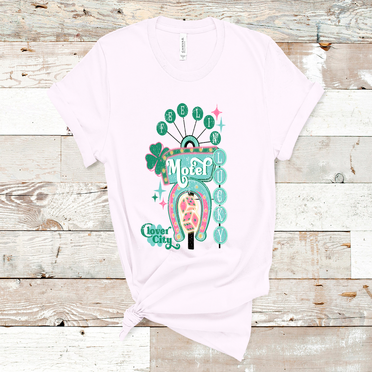 Soft Pink Tee. Graphic of a vintage Neon Sign The word feeling with each letter in green circles at the top with the word lucky down the side with each letter in a turquoise circle. There is a sign in the center with the word motel in it, underneath there is a horse shoe with dice inside at he bottom of the sign. There is also a four leaf clover with the word clover city over it in green.