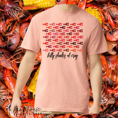A peach tee with a graphic. Graphic is of 50 different crawfish in various shades of red in staggered rows. Under the crawfish there are the words fifty shades of cray in black cursive font.