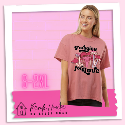 A blonde woman in an oversized mauve HiLo tee with a graphic of a group of mushrooms in various shades of pink. the emushroom in the center has a cap that looks like a heart. In a retro black 70's font it says foraging on top of the mushrooms and for love underneath the mushrooms.