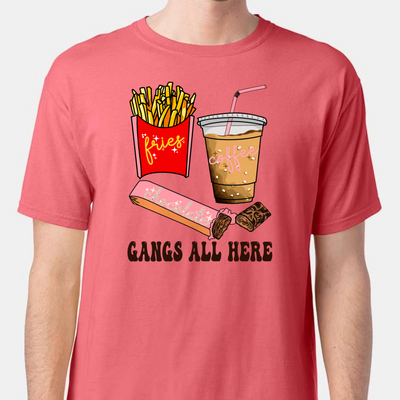 Coral shirt with graphic. Graphic is a container of fries with a yellow cursive font that says fries. A cup of iced coffee with a pink cursive font that says coffee and a chocolate bar witha pink wrapper and a blue cursive font that says chocolate. Under the food there is a black bubble font that says gangs all here.