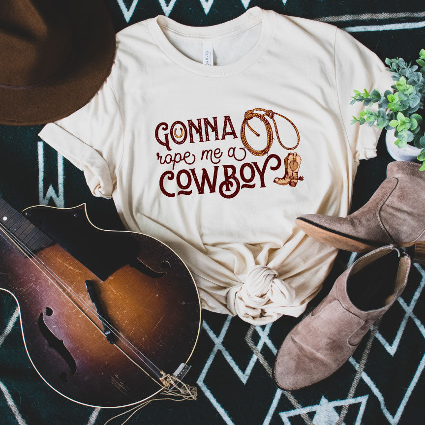 A dust shirt with the words "Gonna Rope me a Cowboy" In a western font. In the O of gonna there is a horseshoe and to the right there is a rope and a cowboy boot.