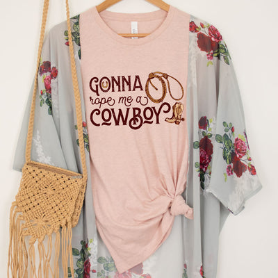 A peach shirt with the words "Gonna Rope me a Cowboy" In a western font. In the O of gonna there is a horseshoe and to the right there is a rope and a cowboy boot.