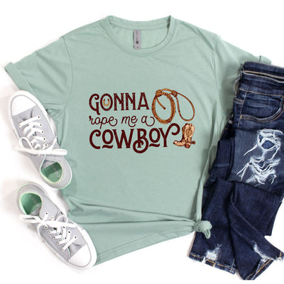 A stonewash green shirt with the words "Gonna Rope me a Cowboy" In a western font. In the O of gonna there is a horseshoe and to the right there is a rope and a cowboy boot.