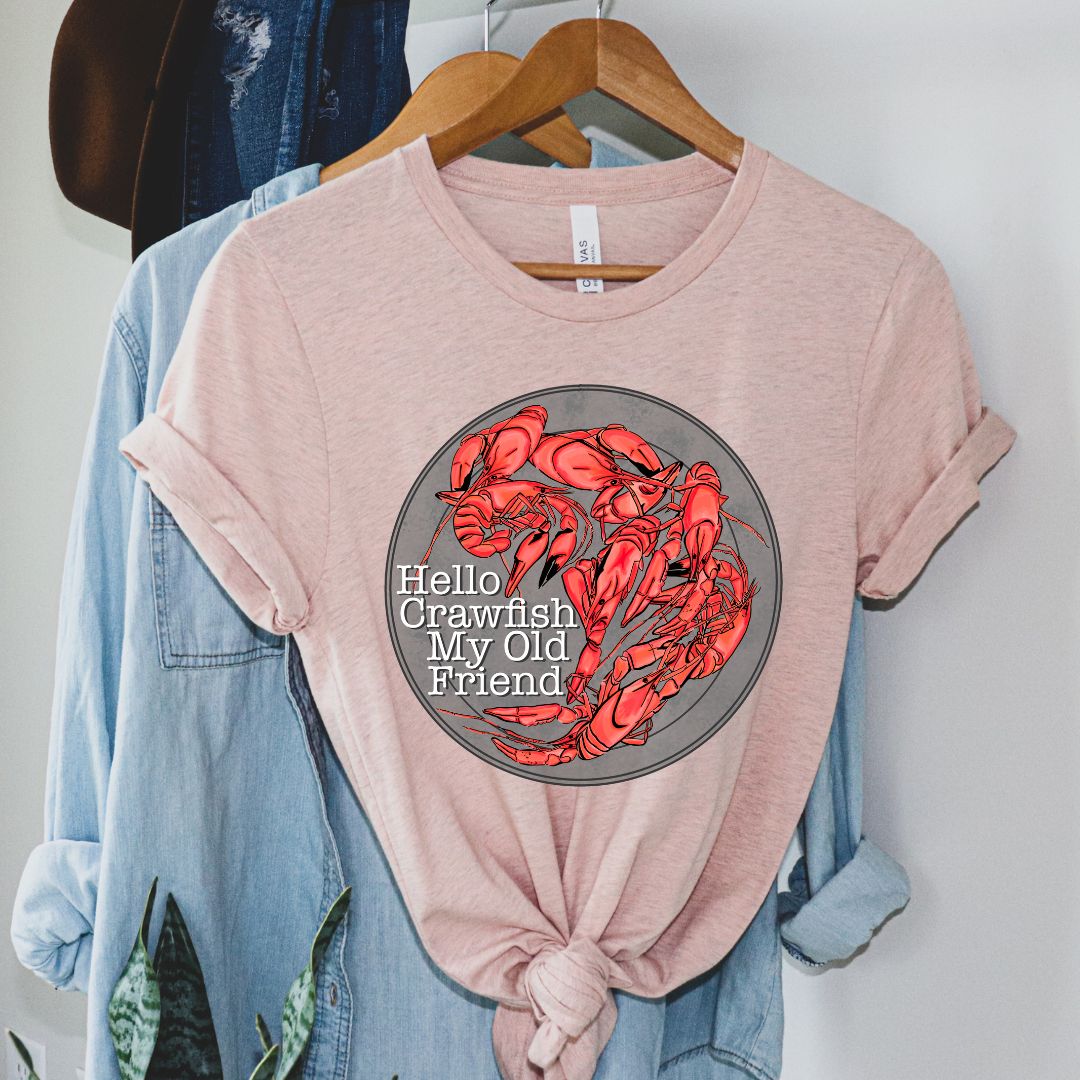 Heather Peach Tee. Graphic is a grey plate full of crawfish and the words "Hello Crawfish My Old Friend" In a white font towards the bottom left of the plate