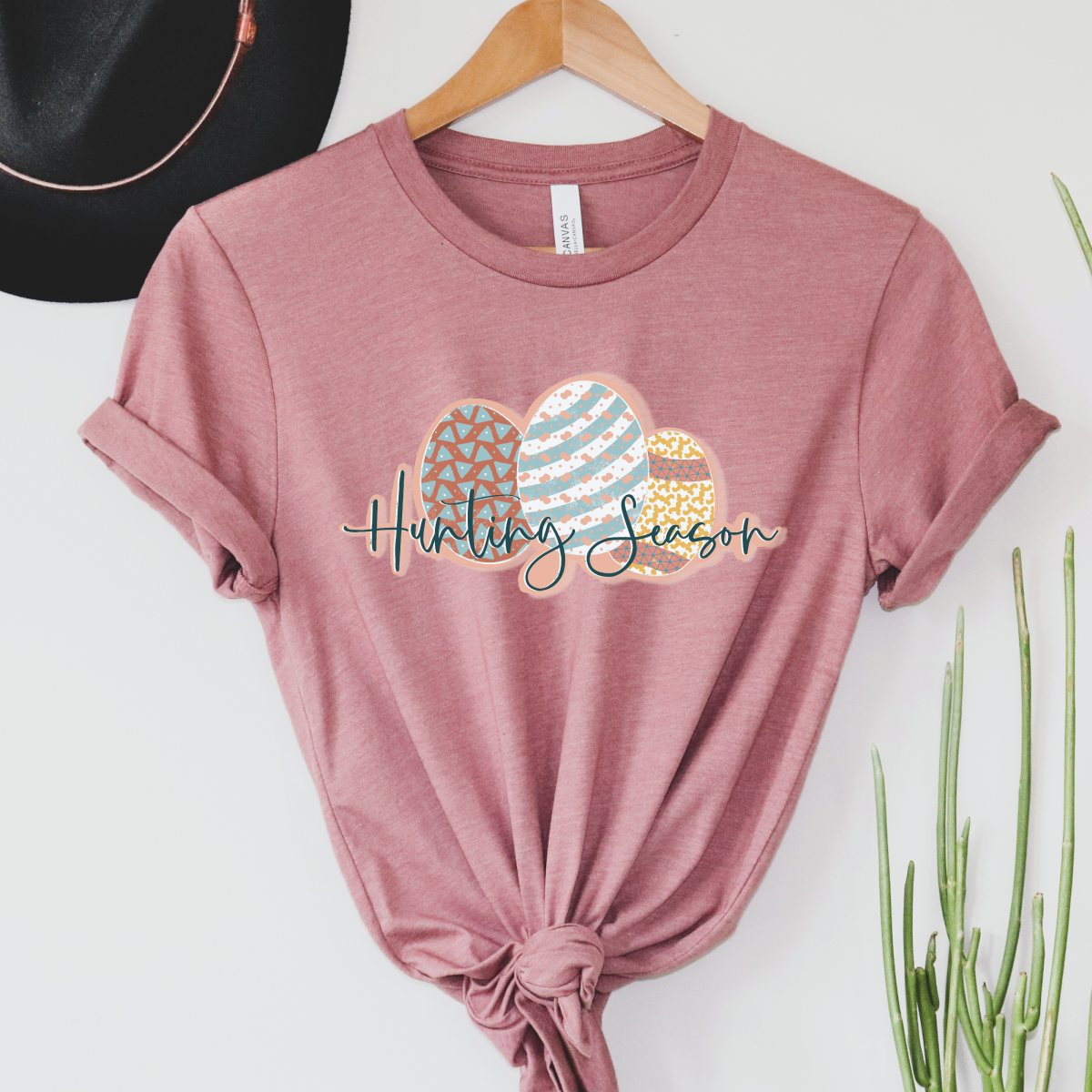 Mauve tee with a graphic of three differently decorated easter eggs and the words "Hunting Season" In a blue cursive font