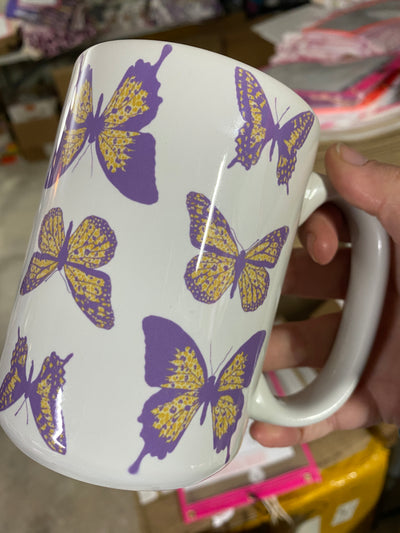 Hand holding a white coffee mug. Coffee Mug has purple and yellow butterflies on it. 
