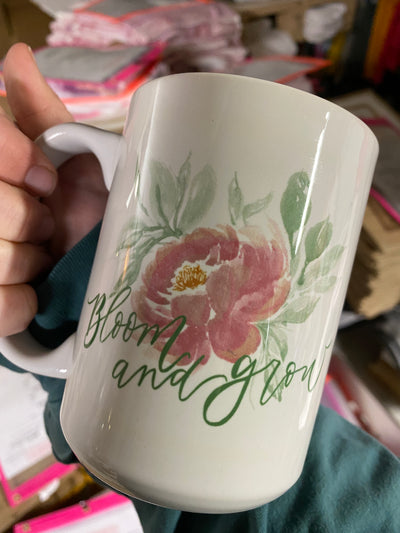 Hand holding a coffee mug. Coffee Mug has a watercolor style pink flower with green leaves and the words Bloom and Grow in green.