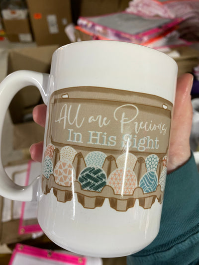 Hand holding a coffee mug. Coffee mug has a carton of decorated eggs and the carton says " all are precious in his sight"