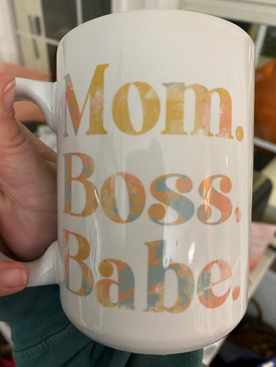 Hand holding a Coffee Mug. Mug says Mom. Boss. Babe. in a blue, peach, yellow watercolor font.