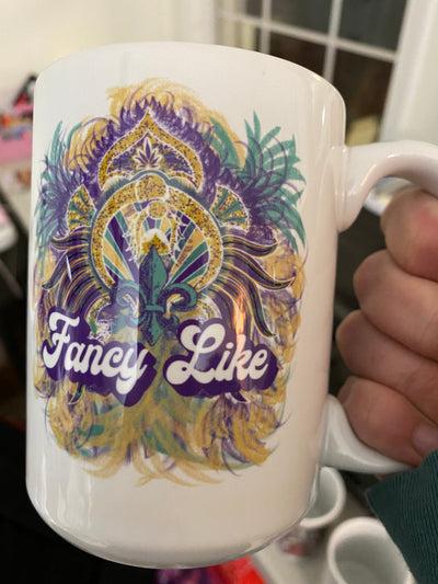Hand Holding a coffee mug. Coffee mug has a decorative Fleur De Lis with feathers and text that says Fancy Like