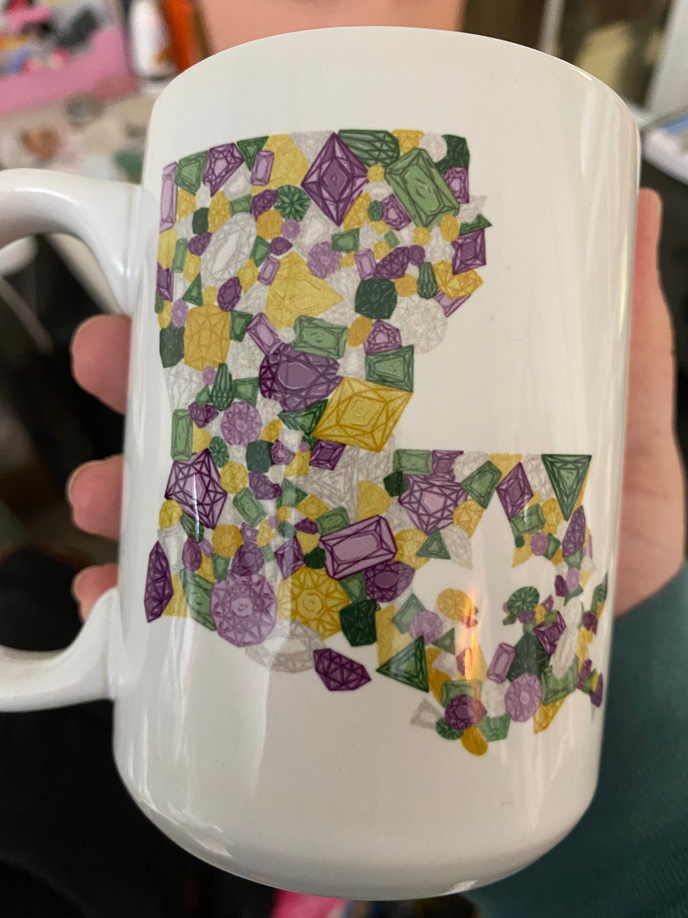 White coffee mug  with a graphic of the state of Louisiana compromised of different shapes and sizes of purple, green, yellow and clear jewels.