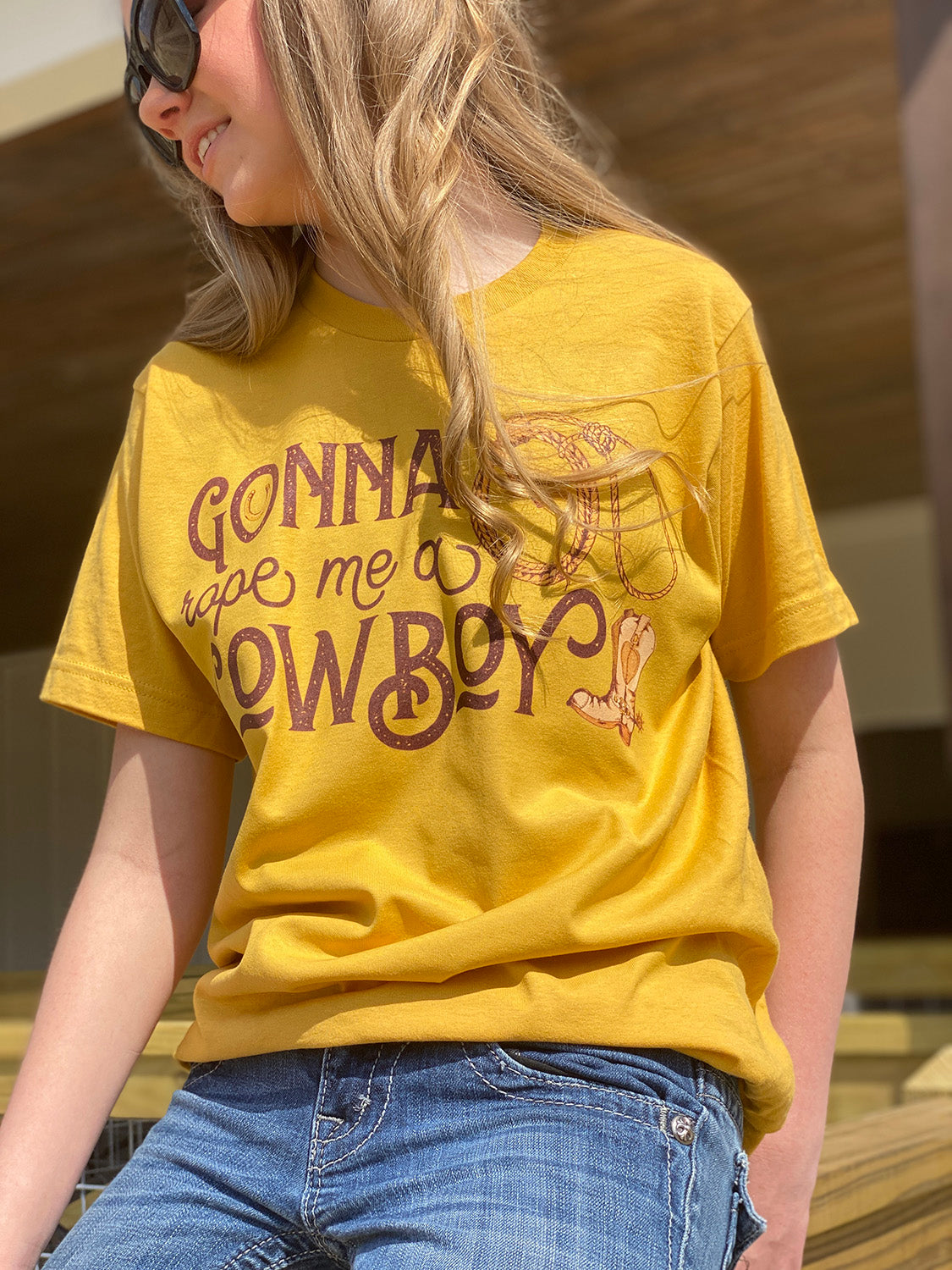A yellow shirt with the words "Gonna Rope me a Cowboy" In a western font. In the O of gonna there is a horseshoe and to the right there is a rope and a cowboy boot.